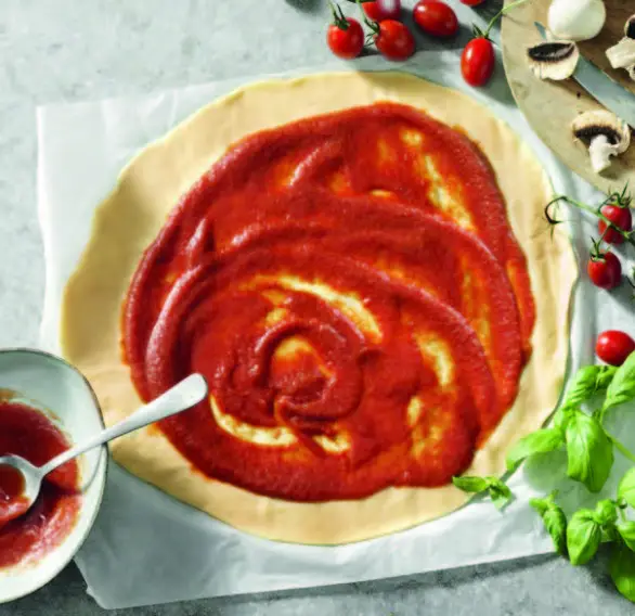 Préparation de pizza avec sauce tomate étalée sur la pâte, accompagnée de tomates cerises, champignons et basilic frais.