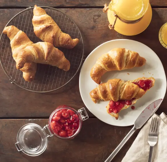 Croissants dorés sur une table en bois, accompagnés de confiture de fraises et de jus d'orange frais.